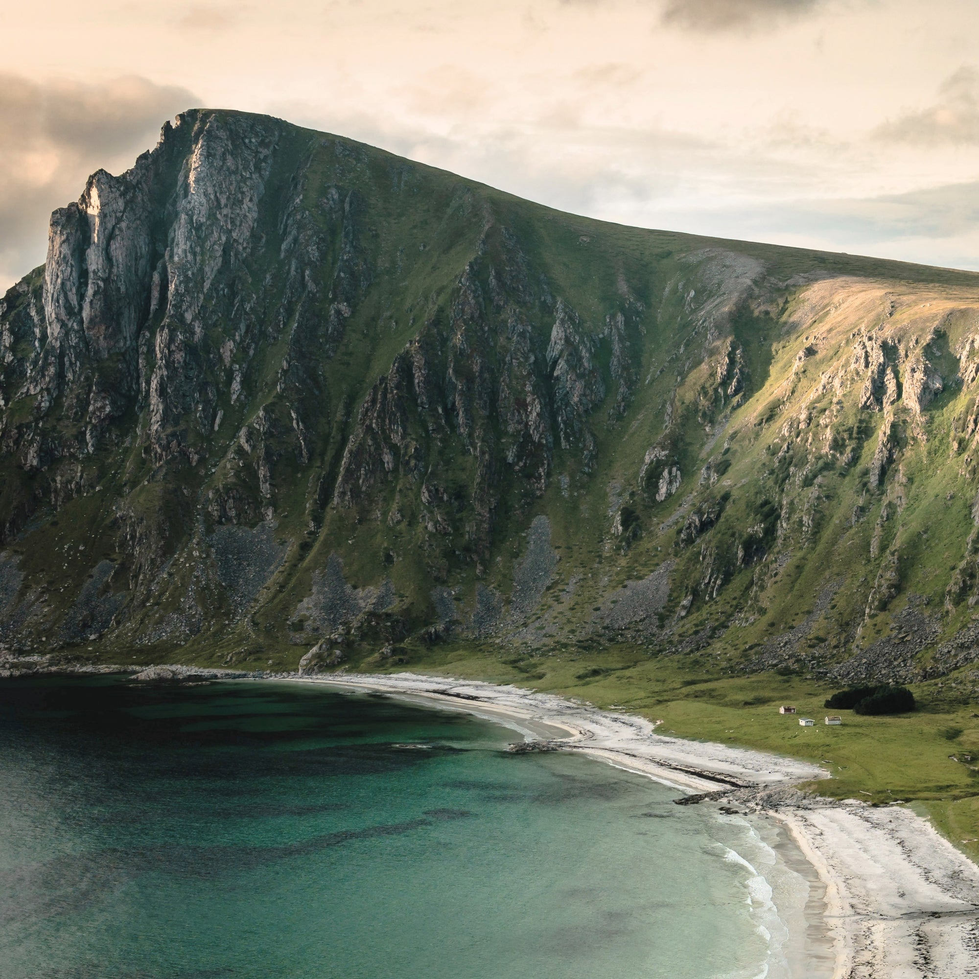 Bratt grønt fjell med grå klipper faller ned mot en hvit sandstrand og turkist hav under delvis skyet himmel
